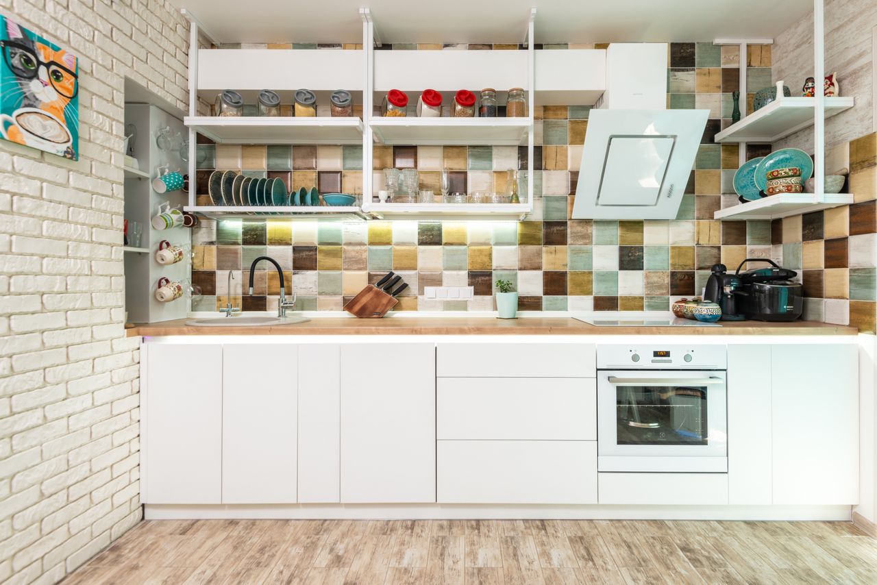 Interior of bright kitchen furnished with white cupboards and shelves with oven decorated with various dishware and utensils with tiled multicolored backsplash wall and brick white wall and wooden floor